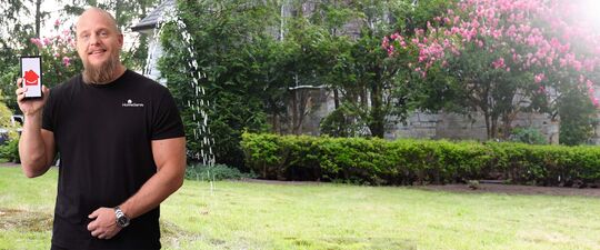 Matt demonstrates home repair subscription plan by holding a smartphone and standing in front of the broken water pipe line in the front yard.