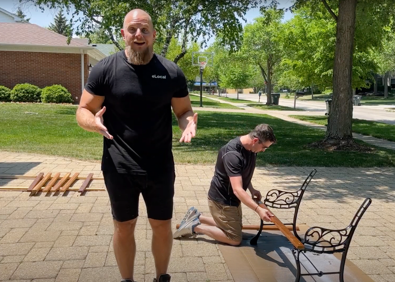 Matt Schmitz stands as he speaks to the unseen video camera while behind him is Eric Rossi kneeling on a residential driveway surrounded by greenery as he works on a partially disassembled garden bench, trees, greenery, grass, lawn, front yard, yard, driveway, paver driveway, pavers, disassembled, bench, garden bench, diy, do it yourself, project, diy project, wood, wooden, wood slats, wood planks, metal, iron, street, residential street, neighborhood street, house, family house