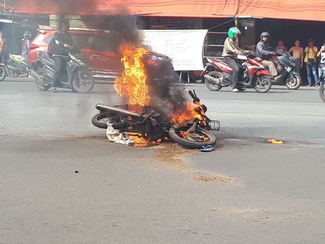 A crashed motorcycle burns in the middle of a city street as other motorcyclists ride past in the background behind the flames and black smoke, motorcycle, motorcycle crash, crash, car crash, auto accident, accident, burning, fire, city street, motorcycles, motorcycle riding, motorcyclists