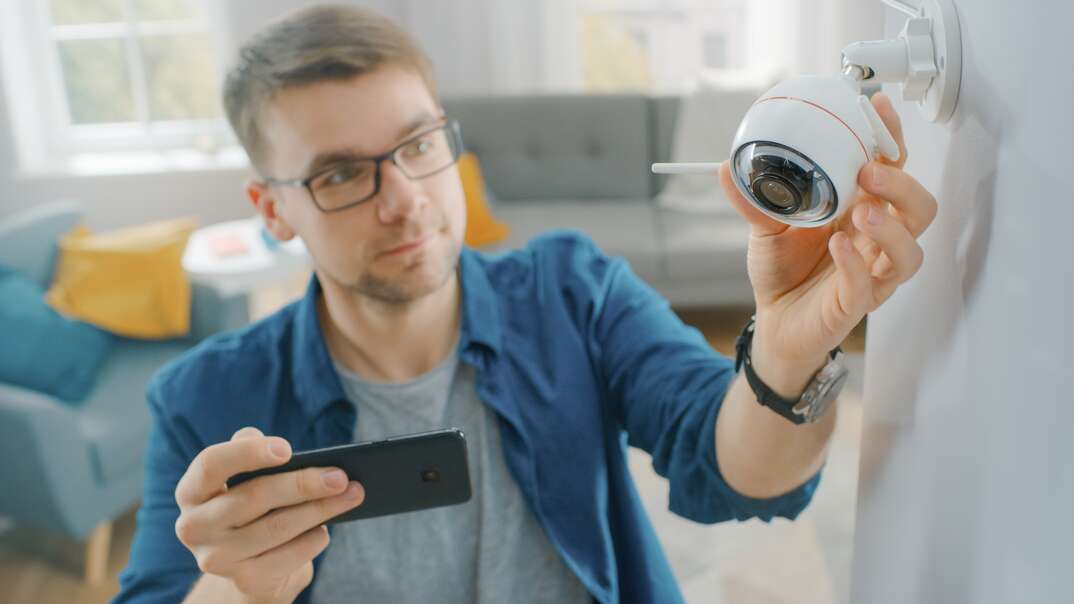 Young Man in Glasses Wearing a Blue Shirt is Adjusting a Modern Wi-Fi Surveillance Camera with Two Antennas on a White Wall at Home. He s Checking the Video Feed on his Smartphone.