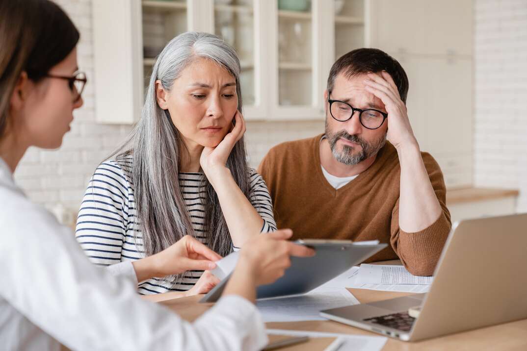 A middle-aged married couple sit at the desk with their financial planner looking distressed as they are shown their retirement income situation, man and woman, man, woman, male, female, husband, wife, husband and wife, married couple, couple, middle-aged man, middle-aged woman, financial planner, retirement planning, financial planning, financial adviser, adviser, accountant, desk, laptop computer, computer, laptop, distressed, nervous, unhappy