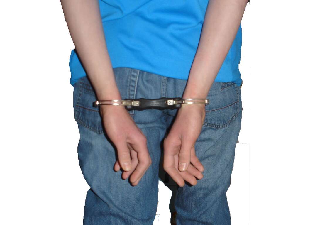 A man wearing blue jeans and a blue shirt is shown from behind with his hands cuffed behind his back while standing against a white background, man, male, perp, perpetrator, criminal, suspect, crime suspect, handcuffed, handcuffs, cuffs, detained, arrested, arrest, detainment, hands behind back, white, white background, male, crime, crime and punishment, jeans, blue jeans, bluejeans, blue shirt, shirt, hands, human, human hands, arms