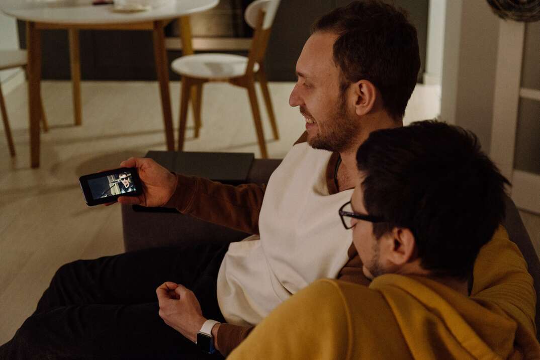 Two adult males sit on a couch together smiling as they look at a photo on a smartphone held by one of the two men, domestic partnership, domestic, partnership, lovers, love, relationship, married, marriage, couple, same-sex couple, same-sex, gay, gay couple, LGBTQ, looking at smartphone, looking at photo, photo, couch, sitting together, sitting, smiling, happy, happiness, in love, togetherness, together