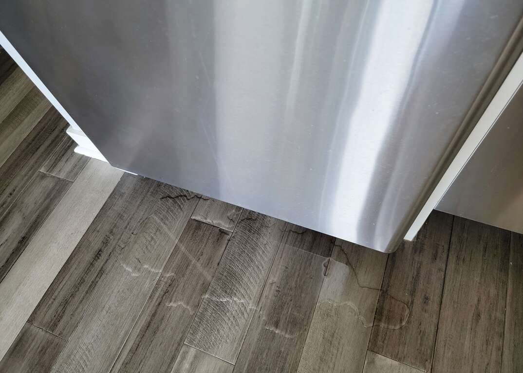 A puddle sits on the gray hardwood floor underneath a stainless steel refrigerator in a modern domestic kitchen