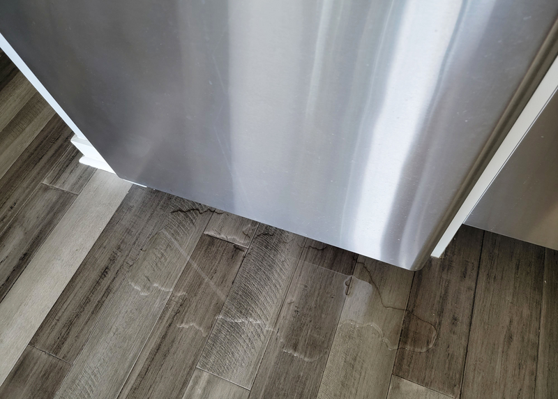 A puddle sits on the gray hardwood floor underneath a stainless steel refrigerator in a modern domestic kitchen, fridge, refrigerator, stainless steel refrigerator, appliance, stainless steel appliance, leak, water leak, water, clear water, puddle, hardwood floors, gray hardwood floors, hardwood, bamboo floors, kitchen, domestic kitchen, kitchen appliance, modern kitchen, leaky fridge, leaky refrigerator, refrigerator leak, fridge leak
