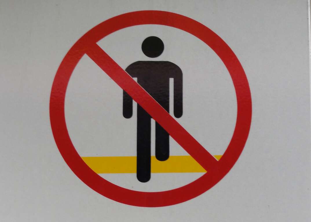 A closeup of a warning sign shows a human figure behind a red circle with a diagonal crossbar through it indicating no trespassing or keep out agains a white background, warning sign, warning symbol, keep out, no trespassing, trespassing, stay back, stay out, keep away, white background, prohibited