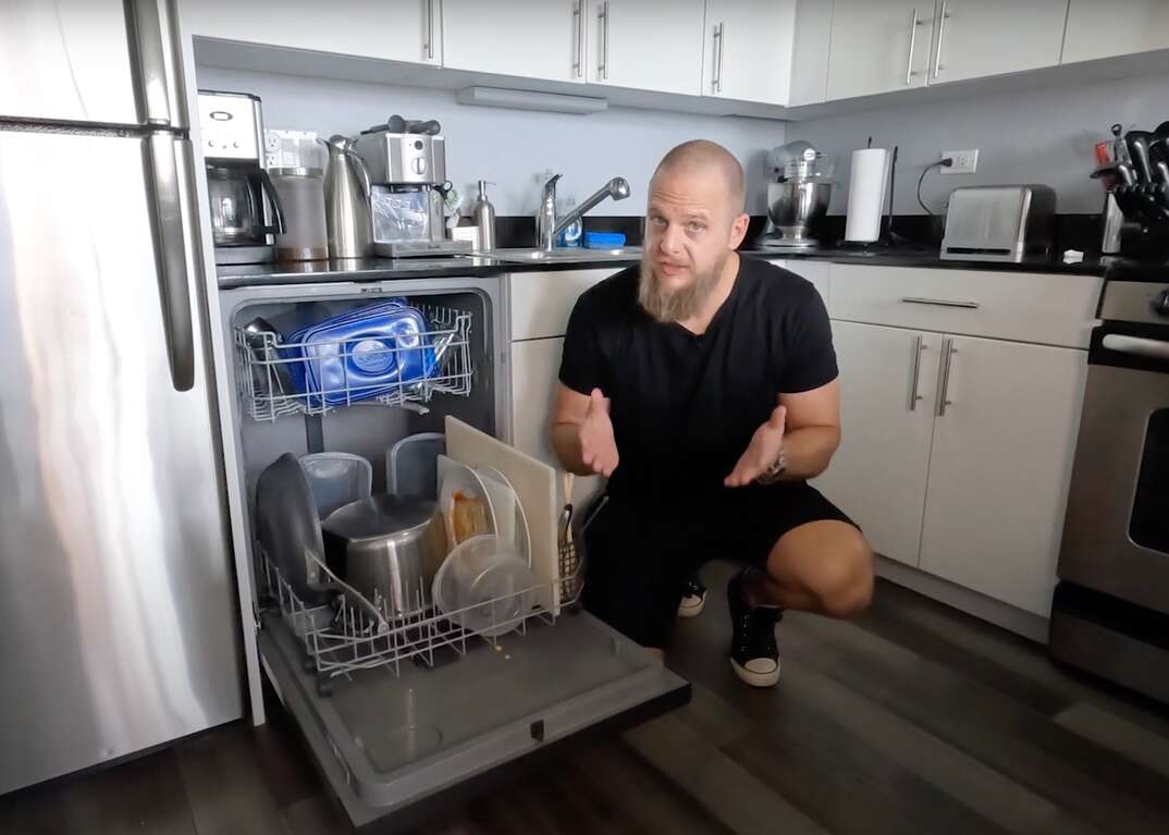 A man wearing a black T shirt and black shorts in a modern kitchen squats in front of a full dishwasher with its door all the way open as he explains how to clean your dishwasher filters, dishwasher, dishwasher filters, kitchen, modern kitchen, Matt Schmitz, Schmitz, Matt, eLocal, stainless steel appliances, appliances, refrigerator, fridge, gray hardwood floors, hardwood floors, hardwood, floors, bamboo floors, oven, stove, kitchen countertop, dishwasher filters, full dishwasher, dishes, cleaning, explaining, video, YouTube video, YouTube