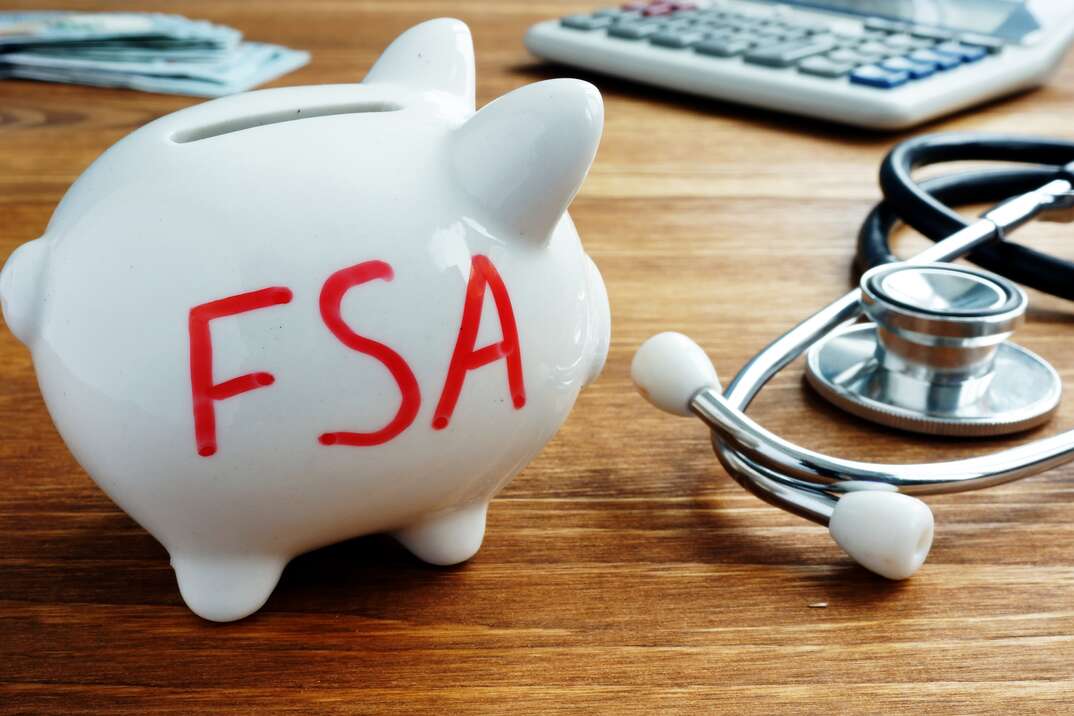 A white piggy bank with red lettering that reads FSA sits atop a brown woodgrain desk next to a stethoscope and a calculator, piggybank, piggy bank, white piggy bank, FSA, flexible spending account, stethoscope, desk, woodgrain desk, calculator, health coverage, health insurance, insurance, coverage, medical coverage, medical insurance