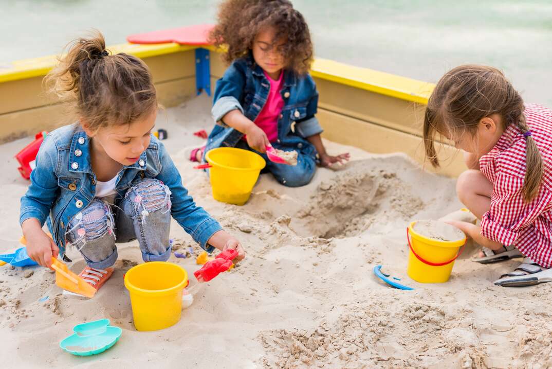 three multiethnic little children playing with plastic scoops and buckets in sandbox at playground