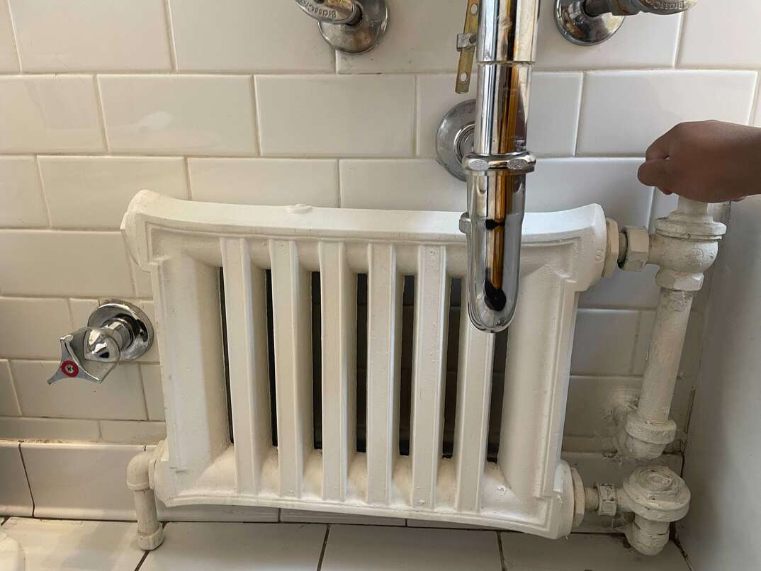 A white radiator stands in a bathroom against a white ceramic tile wall and on a white ceramic tile floor, surrounded by stainless steel pipes.