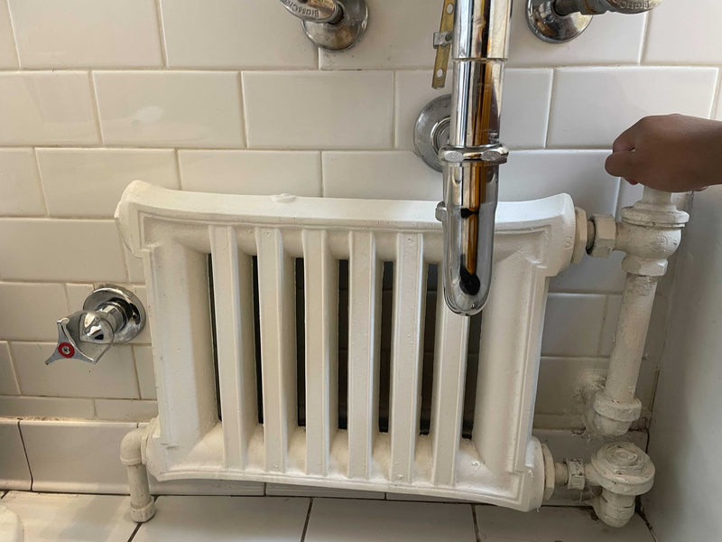 A human hand twits the temperature control knob of a white radiator that stands in a bathroom against a white ceramic tile wall and on a white ceramic tile floor, surrounded by stainless steel pipes, radiator, white radiator, white ceramic tile, ceramic tile, tile, tile walls, bathroom tile, bathroom floor, stainless steel, stainless steel pipes, heater