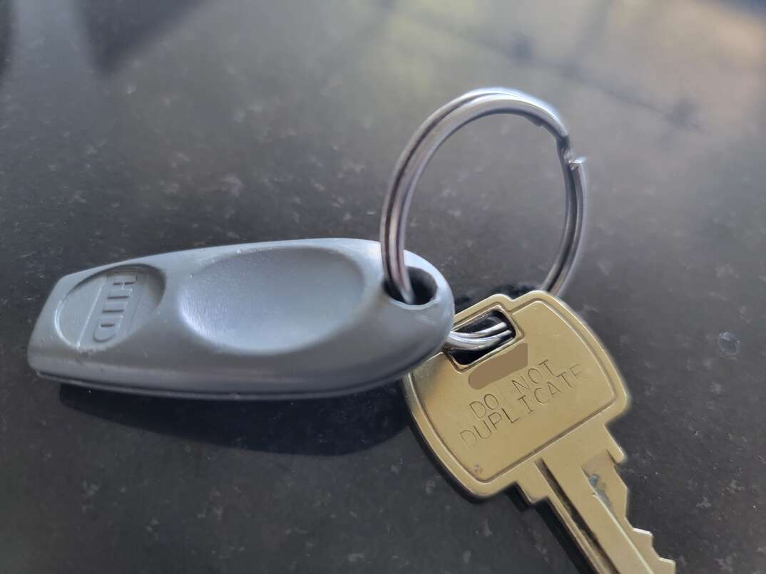 A gray key fob is connected via a keyring to a gold colored house key bearing the words Do Not Duplicate, keyring, gray key fob, key fob, key, fob, house key, gold colored house key, black granite countertop, black granite, granite, black