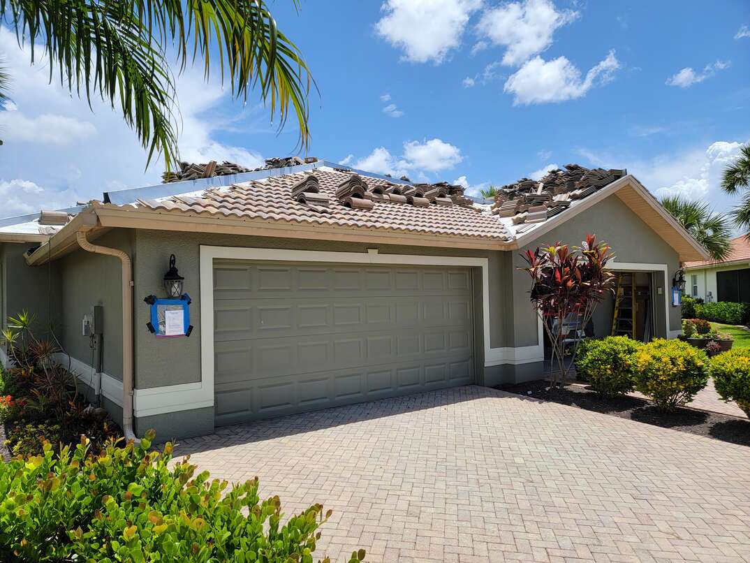 A villa in Florida gets its Spanish tile roof replaced above the garage as a palm tree hangs into the frame and the home sits against a blue sky with white clouds, Spanish tile, Spanish, tile, Spanish tile roof, tile roof, roof, Spanish roof, driveway, blue sky, clouds, greenery, shrubs, palm tree, tree, palm