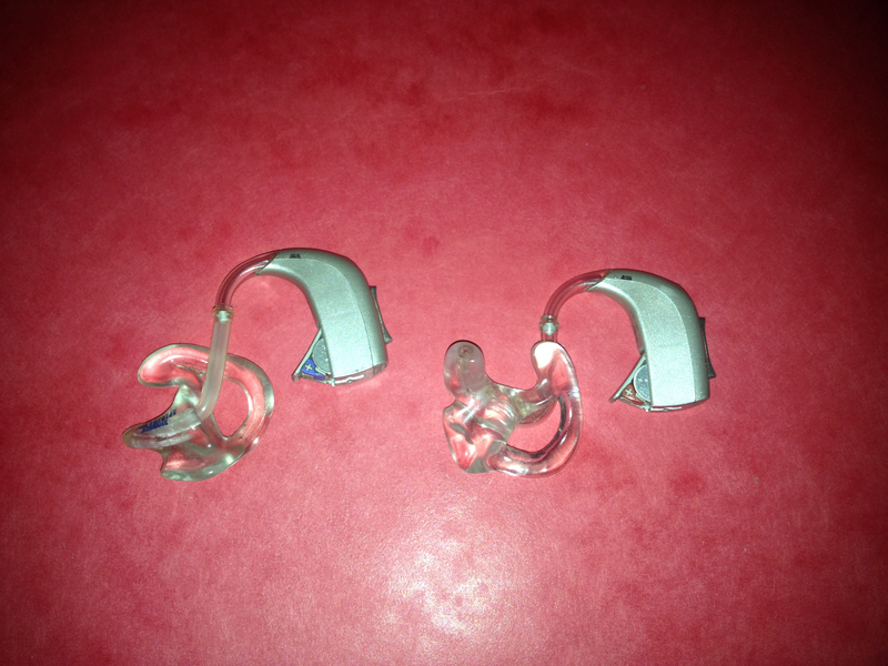 A pair of hearing aid devices sit on a red surface, pair of hearing aid devices, pair, hearing aid, hearing aids, device, devices, hearing, aid, ears, human ears, red, red surface, red table, health, healthcare, health care, technology