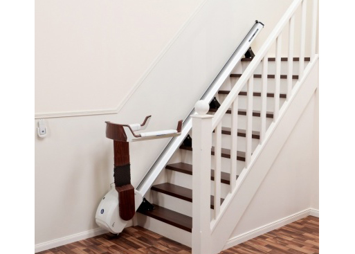 A chair lift device is shown installed along the wall of a residential home staircase against a white wall and a white banister with brown wood floors, chair lift, stair lift, stairs, steps, house, home, accessible, accessibility, handicapped, disabled, device, technology, wood floor, brown wood floor, white walls, white background