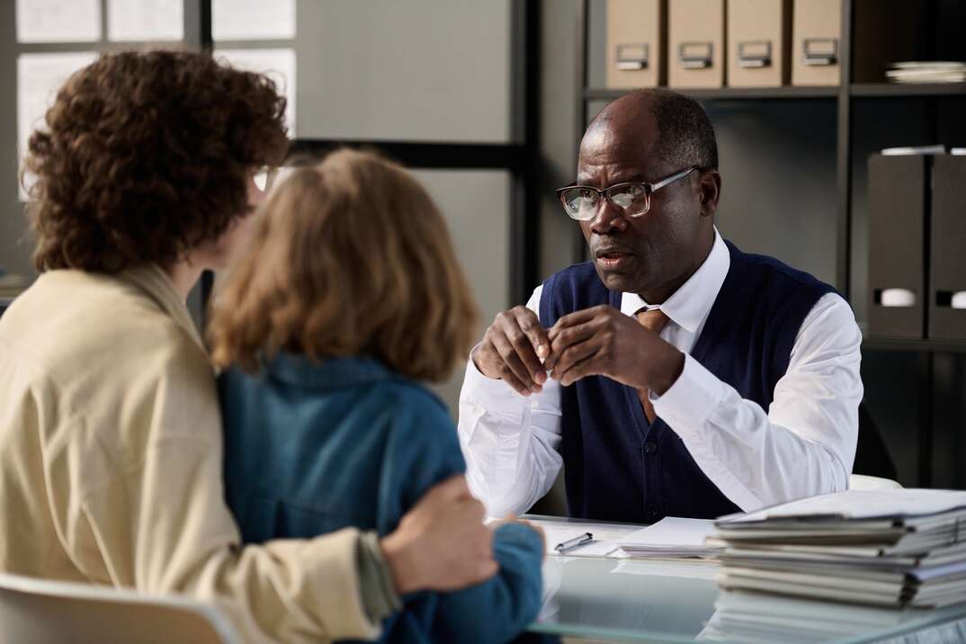 A lawyer sits at his desk in front of shelves of legal books and speaks to two clients seated at the other side of the desk, lawyer, attorney, male attorney, male lawyer, law, law office, lawyer's desk, lawyer's office, clients, law books, legal books, books, binders, desk, legal, legal advice