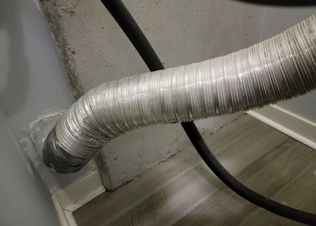 An aluminum dryer vent tube leads from a clothes washer to the dryer vent in a gray wall, metallic, aluminum, gray, silver, tube, dryer tube, dryer hose, clothes dryer, gray hardwood floor, white floorboard, hardwood floor, floorboard, black hose