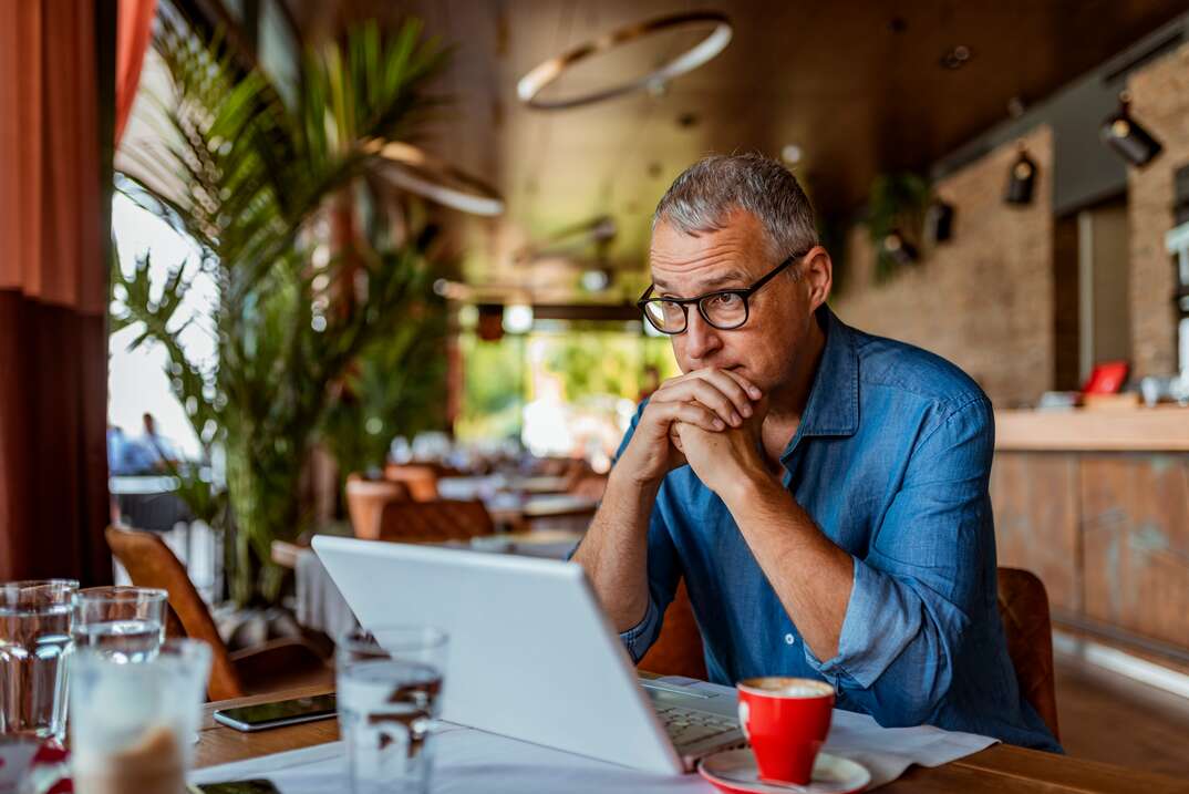 An older man wearing glasses and a blue shirt sits at a table with his chin resting against his clasped hands as he stares at the screen of a laptop computer looking distressed, older man, glasses, eyeglasses, wearing glasses, blue shirt, table, kitchen table, desk, laptop computer, laptop, computer, plants, houseplants, distressed, worried, nervous, at home, home, house, bills, paying bills