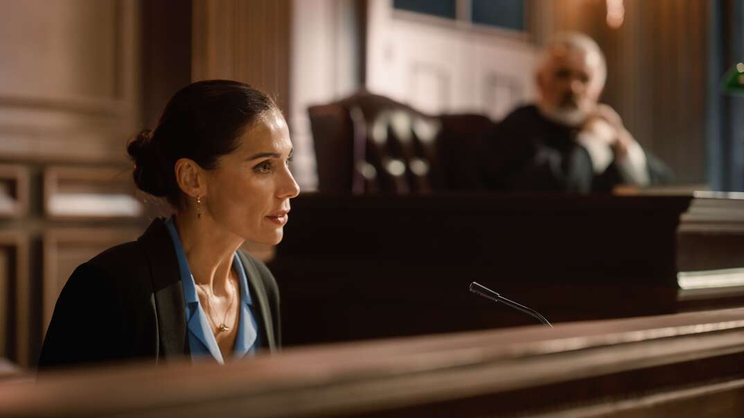 A woman in business attire sits on the witness stand looking intently in the foreground as a bearded male judge looks on from the bench in the blurred background, female witness on the stand, on the stand, witness, expert witness, witness testimony, cross-examination, testimony, court, courtroom, legal, law, attorney, judge, male judge, bearded, beard, bench judge's bench, witness stand, the stand