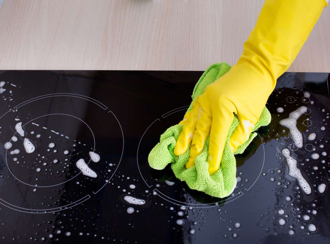 Close up of a hand with yellow protective gloves cleaning induction cooktop with green microfiber cloth