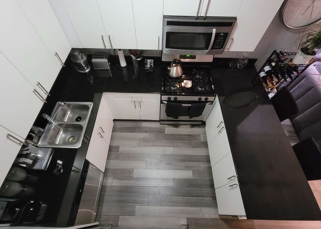 Overhead view of a modern kitchen with white cabinets, black granite countertops, gray hardwood floors and stainless steel appliances with an open floorpan leading to a living room with a wine rack and a gray couch.