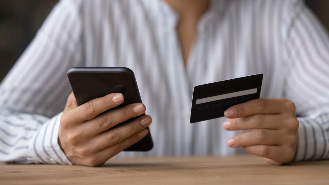 A woman wearing a white long sleeve button down shirt holds a credit card in one hand and a smartphone in the other