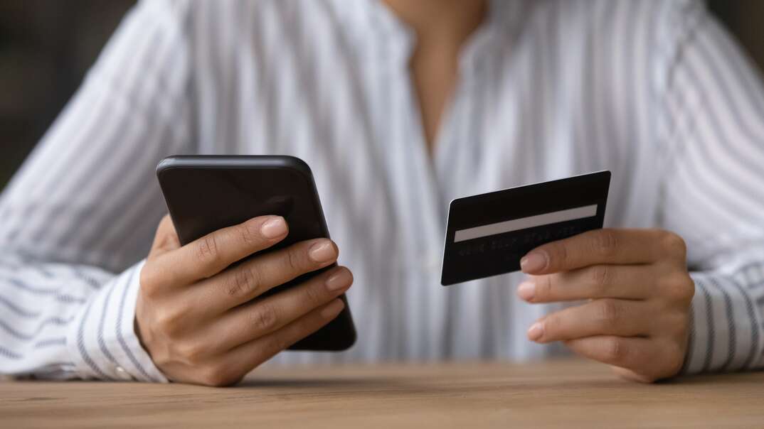 A woman wearing a white long sleeve button down shirt holds a credit card in one hand and a smartphone in the other, woman wearing white long sleeve button down shirt, long sleeves, button down, white shirt, credit card, charge card, card, smartphone, cell phone, phone, mobile phone, mobile, phone scam, scam, crime, identity theft, financial, financial crime, victim