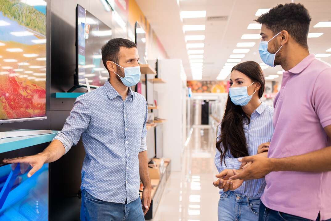 A COVID masked salesman in a brightly lit electronics store shows a masked male and female couple of shoppers a flatscreen TV on the shelf