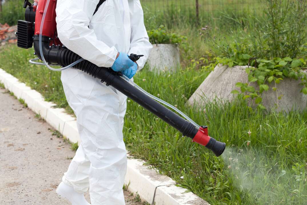 A professional exterminator in a protective white hazmat suit and a red and black sprayer pack on his back sprays a green grassy outdoor area for mosquitoes