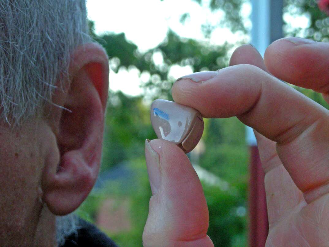 An elderly man with gray hair holds a hearing aid between his thumb and forefinger just a few inches from his ear, elderly man, hearing aid, hearing, health device, disabled, disability, handicap, handicapped, differently abled, ear, man's ear, elderly man's ear, gray hair, elderly man with gray hair, man with gray hair, human hand, thumb, fingers, aid, device, technology, health, trees, greenery, outside, outdoors