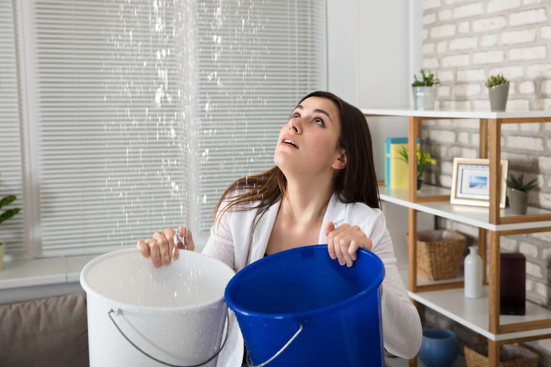 Worried Woman Holding Two Buckets While Water Droplets Leaking From Ceiling