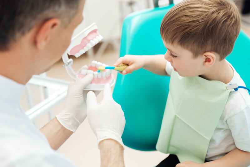 Dentist teaching cute little boy bruching teeth with toothbrush in dental clinic