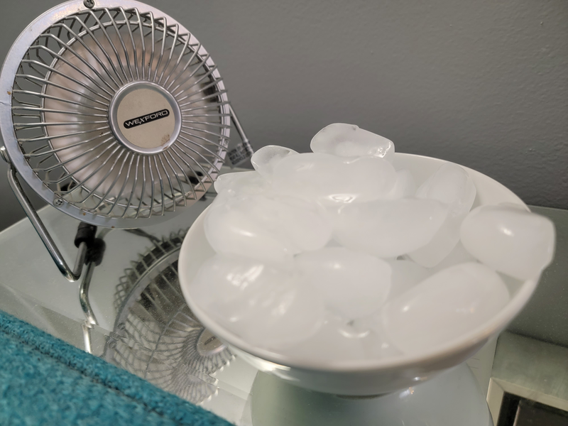 A small electric fan sits atop a mirrored accent table blowing across a white ceramic bowl full of ice, fan, electric fan, metal fan, ice, bowl of ice, white ceramic bowl, bowl, blue couch, couch