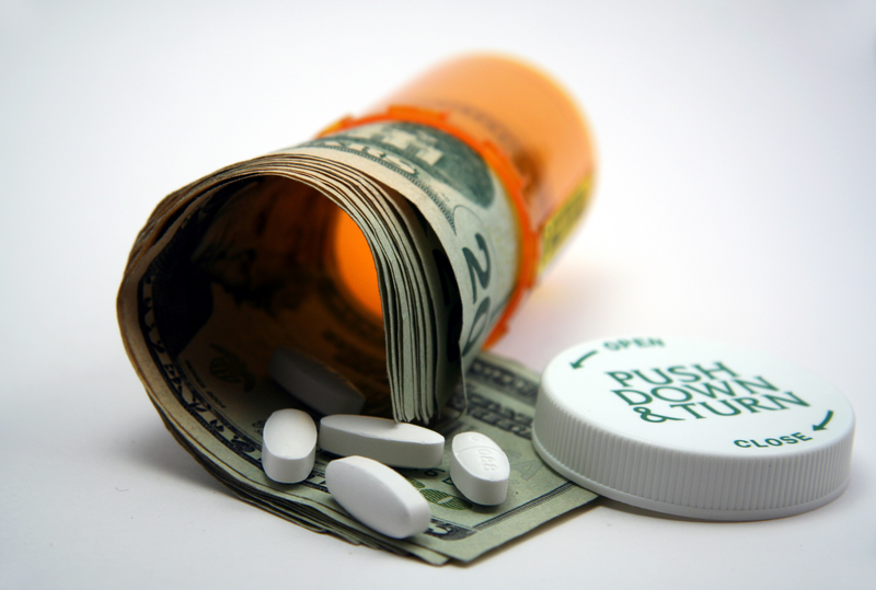 A prescription pill bottle is tipped over agains a white background with the lid off and has white pills and twenty dollar bills spilling out of it, pill bottle, pills, prescription pill bottle, prescription pills, prescription, copay, health insurance, insurance, health coverage, coverage, health care, health, twenty-dollar bills, money, cash, white background