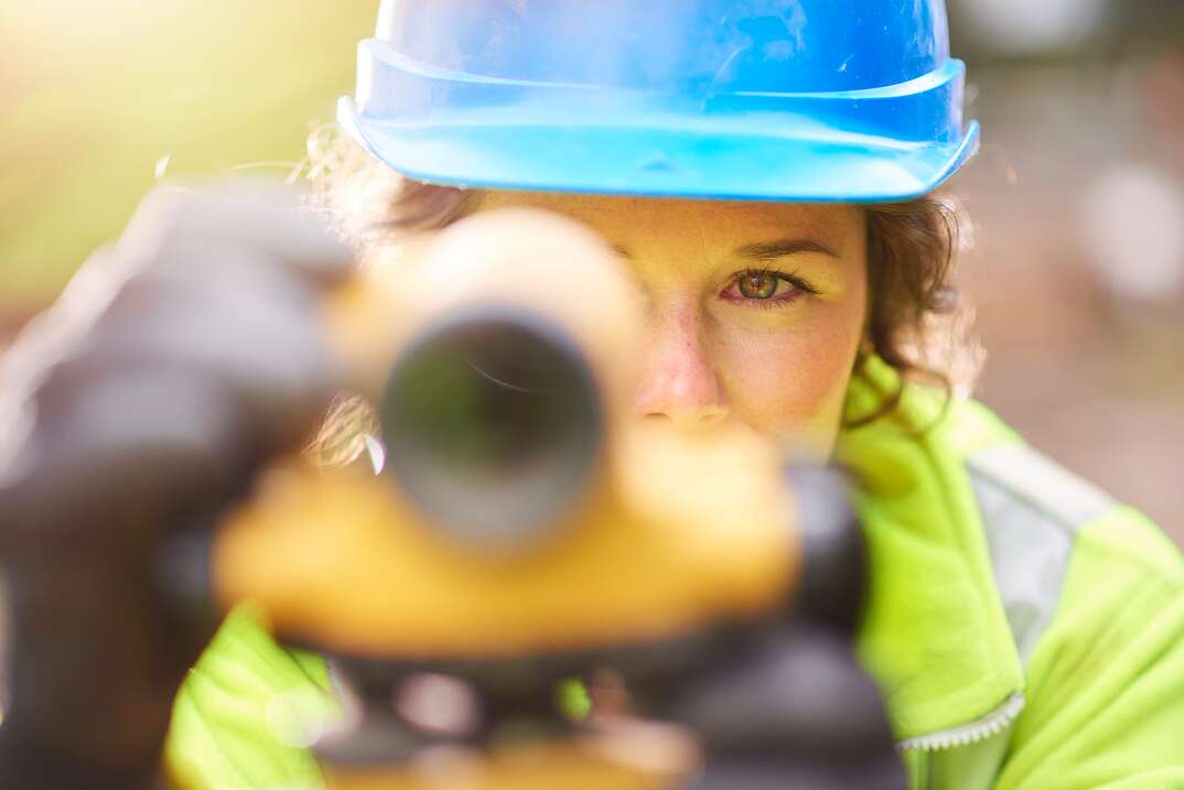 A female technician wearing a yellow safety jacket and a blue work helmet and gloves looks through the viewfinder of the instrument she is using to survey land or measure property lines, land surveyor, surveyor, property lines, measuring property lines, woman, female, female worker, female technician, female surveyor, blue work helmet, work helmet, yellow safety jacket, safety jacket, jacket, gloves, wearing gloves, viewfinder, instrument, instrument of measurement, measurement