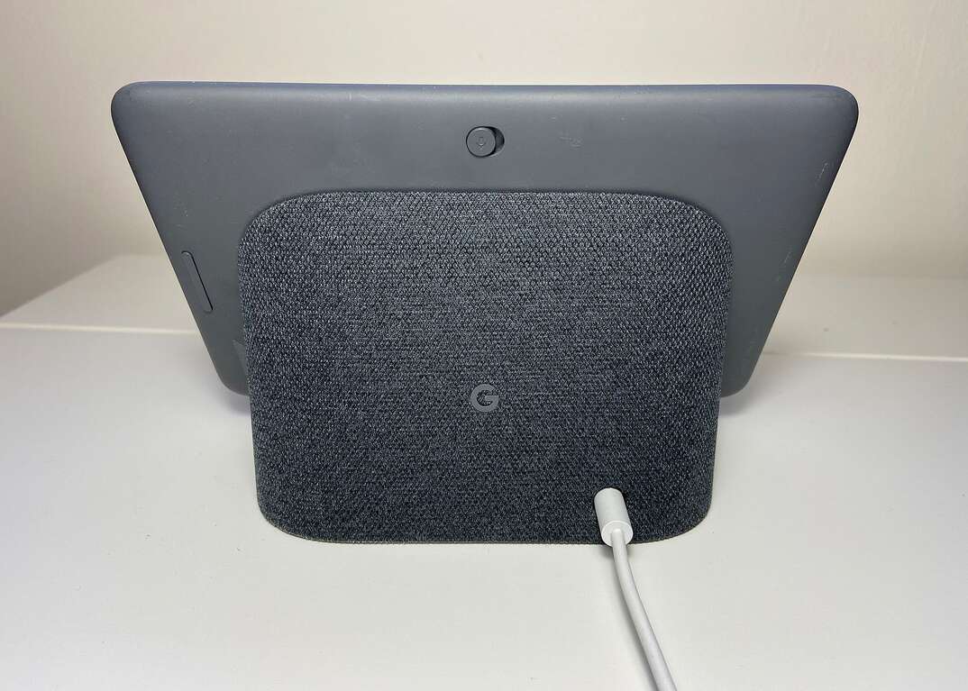 Backside view of a gray Google Nest Hub tablet resting on a white table with a cord coming out of it.