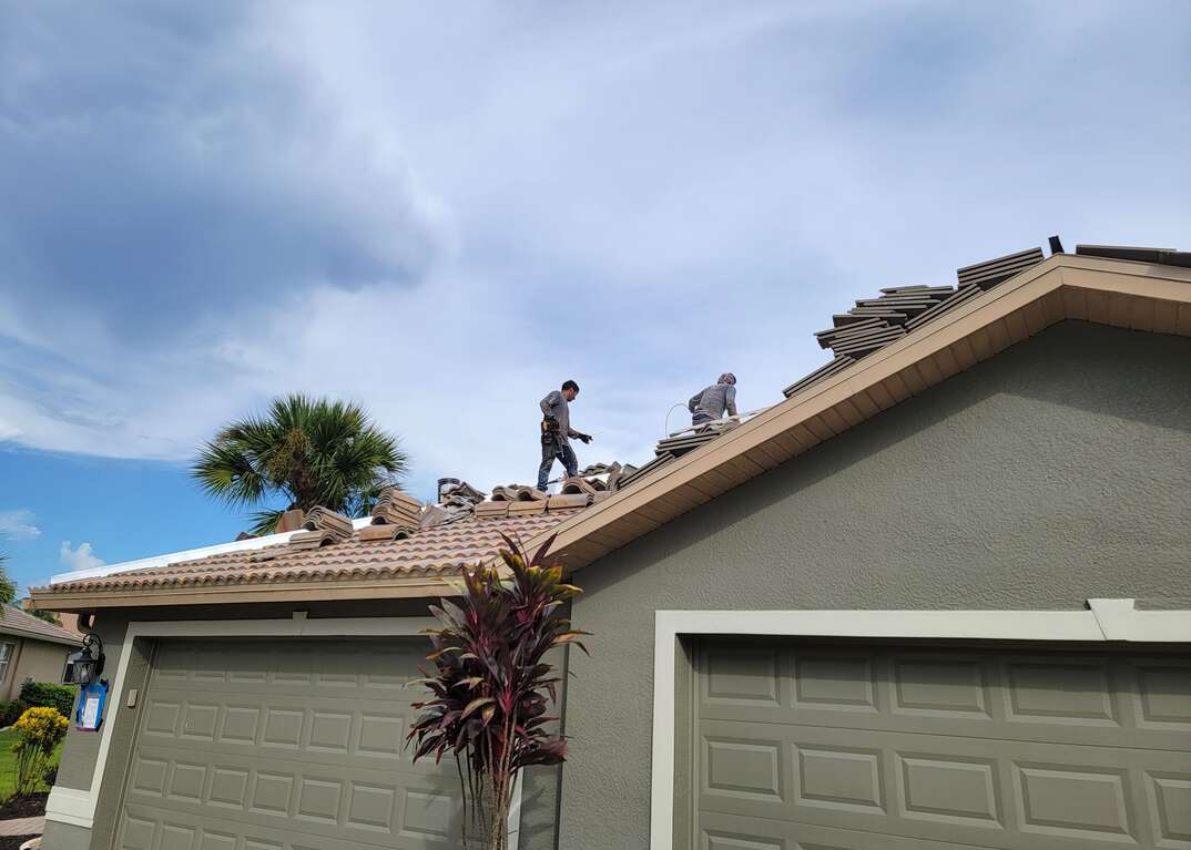 Roofers work atop the roof of a villa installing concrete Spanish tiles against a cloudy sky with a palm tree in the background