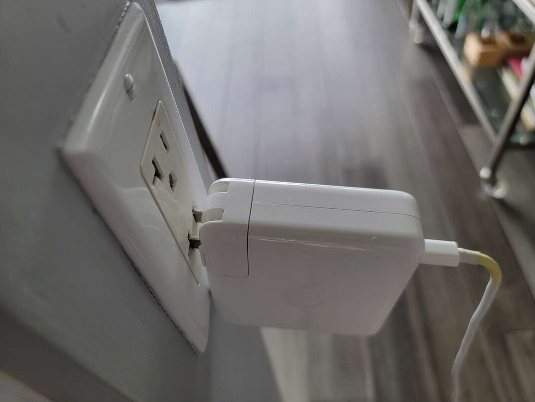 A white Apple computer charger is loosely plugged into a wall socket with a gray hardwood floor and a metal cocktail cart in the background