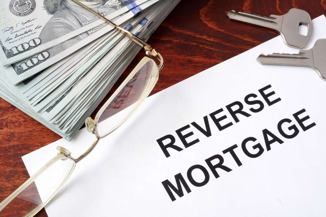 A reverse mortgage document sits on a wood desktop along with a set of house keys and reading glasses and a stack of cash