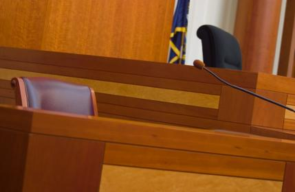 A closeup view in a courtroom shows an empty chair  and a microphone on the witness stand as well as an empty chair and a flag on the judge bench behind it, witness stand, witness, stand, courtroom, court, court case, legal, trial, judge, bench, judge's bench, microphone, flag, wood, wooden, woodgrain