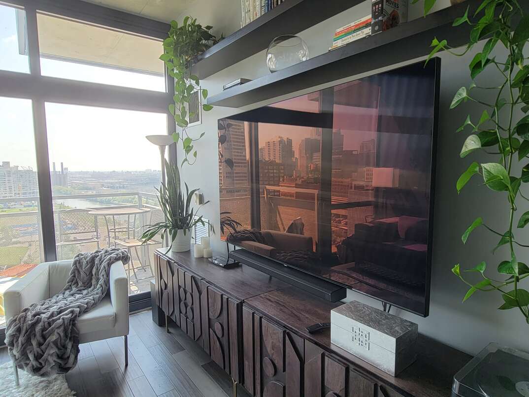 TV, flatscreen TV, television, flatscreen television, condo, sliding glass door, window, hardwood floor, wood floor, gray floor, white chair, chair, armchair, blanket, throw blanket, gray, gray blanket, buffet, entertainment center, brown furniture, reflection, skyline, urban skyline, plants, green plants, hanging plants, floating shelves, bookshelves