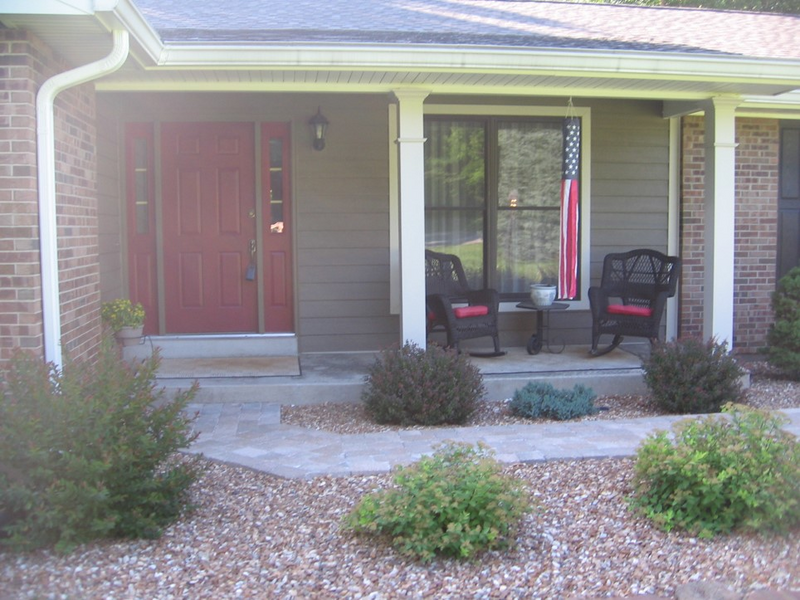The front of a house is shown with a red front door and shrubs and pebble landscaping and an American flag, front of house, flag, American flag, pebbles, shrubs, shrubbery, door, red door, front door