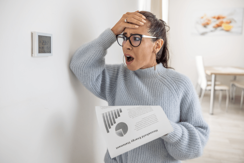 Woman cannot believe her own eyes the sum on her energy bill looking shocked at the thermostat on the wall next to her 