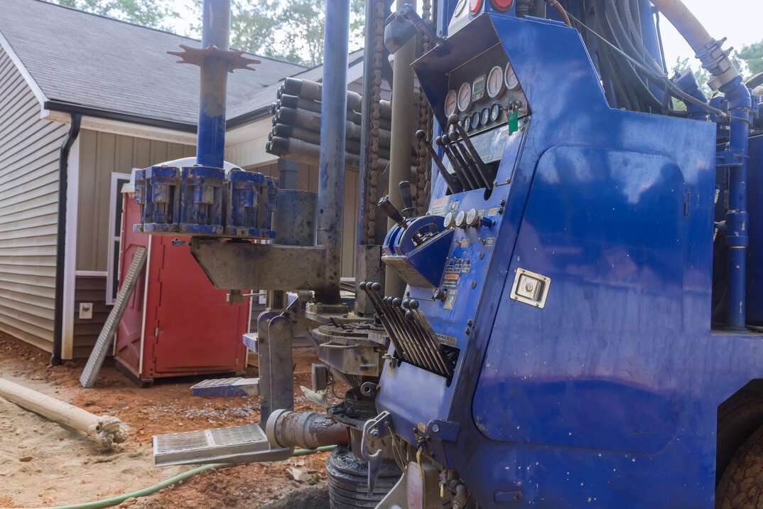 A blue drilling truck sits in front of a house on a dirt lot as it prepares to dig a residential water well