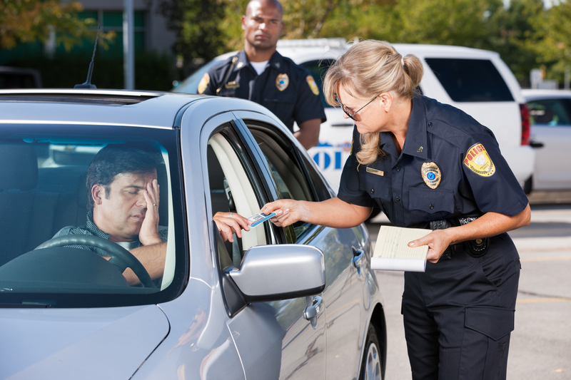 A female police officer standing outside a stopped car occupied by a man who has an exasperated expression as the cop hands his driver license back to him while a male police officer stands behind the vehicle and observes the interaction, car, stopped car, pulled over, traffic stop, police officer, female police officer, woman, woman police officer, male police officer, man police officer, officer, cop, police, uniformed officers, male driver, driver, vehicle, police vehicle, police car, traffic violation