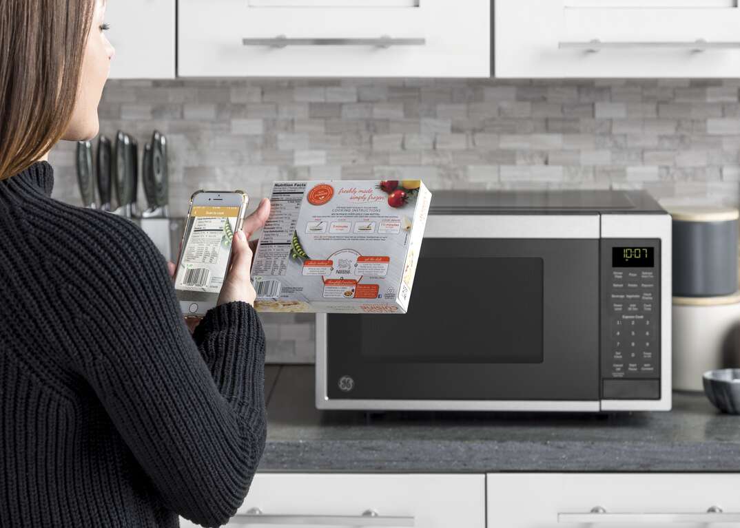 A woman holds her smartphone up to a food package while preparing to cook using a GE brand smart microwave sitting on the countertop in a modern domestic kitchen