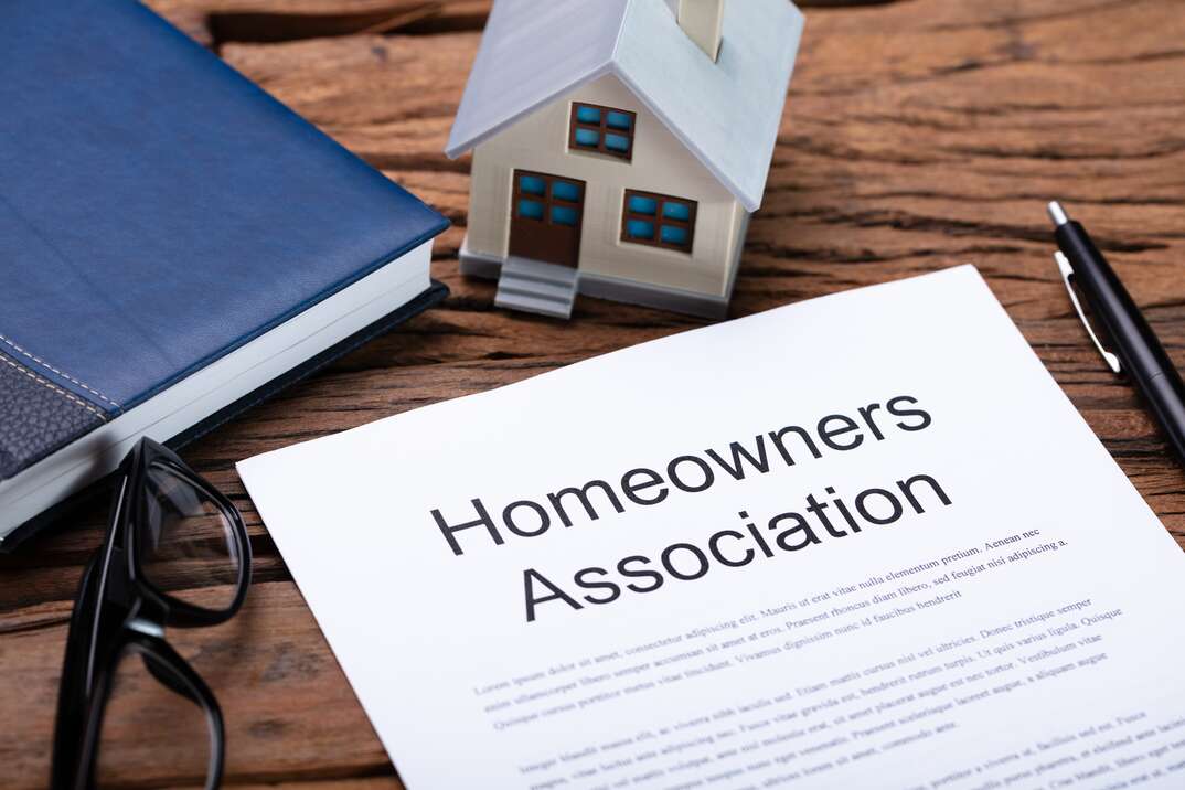 A document presumably containing the rules and regulations and bylaws of a Homeowners Association sits on top of a woodgrain desk surrounded by a pair of eyeglasses and a blue colored book and a miniature house model and a pen, homeowners association, HOA, HOA rules and regulations, rules and regulations, bylaws, HOA bylaws, homeowners association bylaws, desk, woodgrain, woodgrain desk, eyeglasses, glasses, reading glasses, book, binder, blue book, blue binder, miniature house, model house, house model, house, home, model, miniature, pen, inkpen, document, legal document