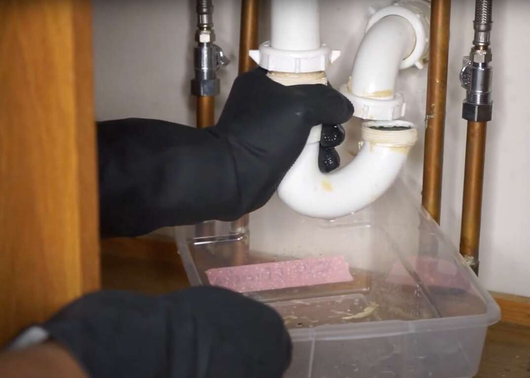 Hands wearing black rubber gloves unscrew the P-trap from underneath a bathroom sink, black rubber gloves, rubber gloves, under sink, sink, pipes, plastic pipes, copper pipes, sink cabinet, bucket plastic bucket