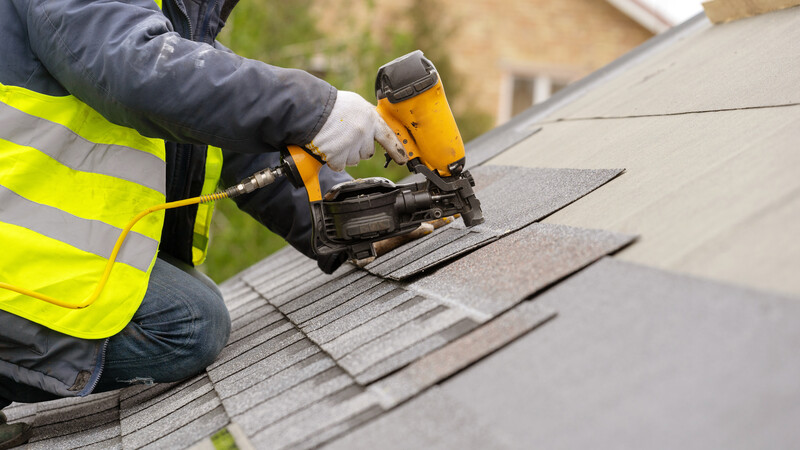 Unrecognizable roofer worker in special protective work wear and gloves, using air or pneumatic nail gun and installing asphalt or bitumen shingle on top of the new roof under construction residential building