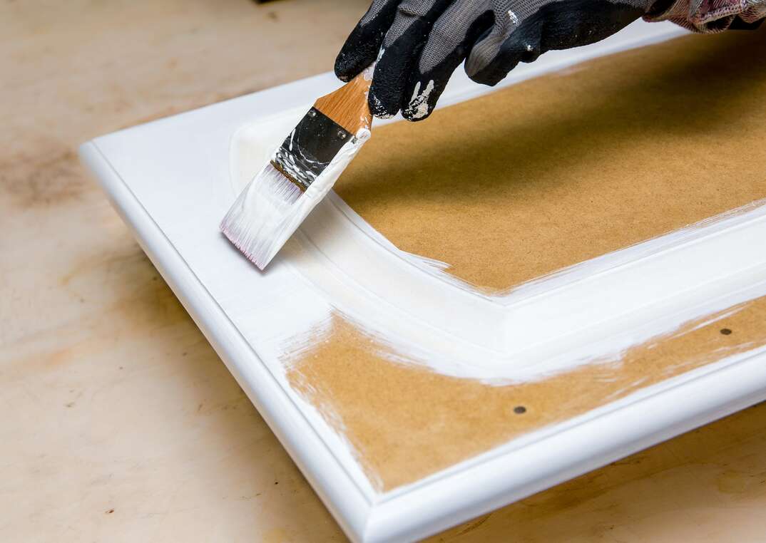 Repainting kitchen cabinet doors with white chalk paint indoors at home. Giving old kitchen new look concept. Hand holding a paint brush tool with paint against old cupboard door.